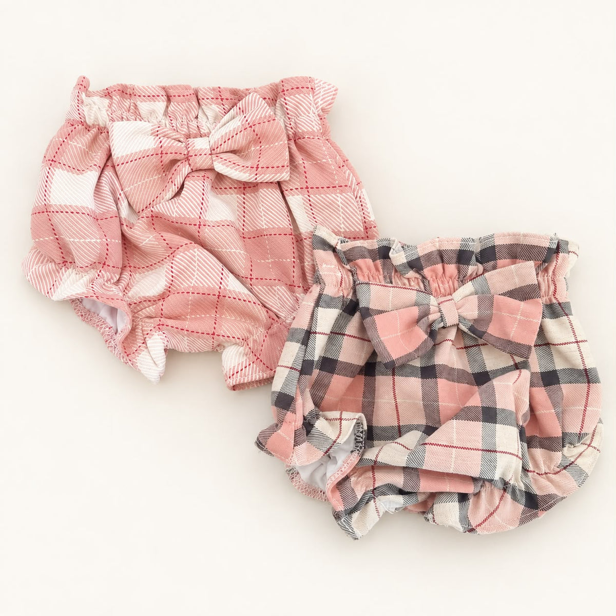 Dois tapa-fraldas para bebé com estampado xadrez em tons de rosa, um com laço rosa e branco e outro com laço rosa, cinzento e branco.
